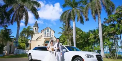 A bride and groom posing with a white luxury SUV in front of a tropical venue with palm trees.