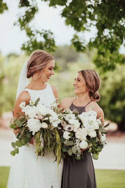 A bride and a bridesmaid smiling while holding large white wedding bouquets with greenery outdoors.