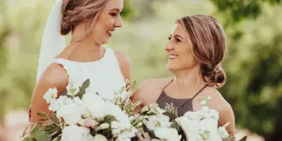A bride and a bridesmaid smiling while holding large white wedding bouquets with greenery outdoors.