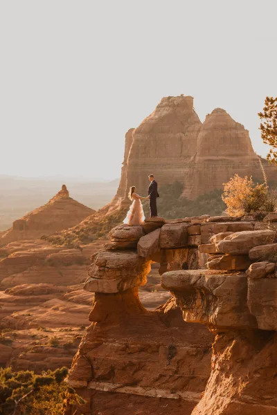 A bride and groom standing on a sandstone rock formation in a desert landscape at sunset.