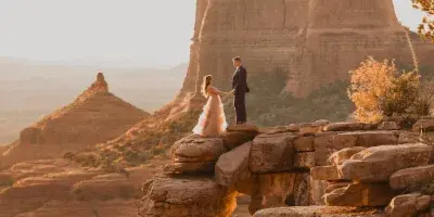 A bride and groom standing on a sandstone rock formation in a desert landscape at sunset.