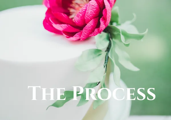 A close-up of a white cake decorated with a pink flower and green leaves.