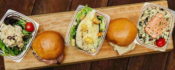 A wooden board holding several catering box meals, including a salad, a sandwich, and a pasta dish with salmon.
