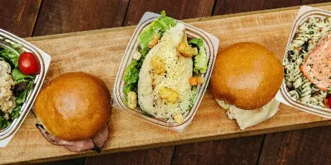 A wooden board holding several catering box meals, including a salad, a sandwich, and a pasta dish with salmon.