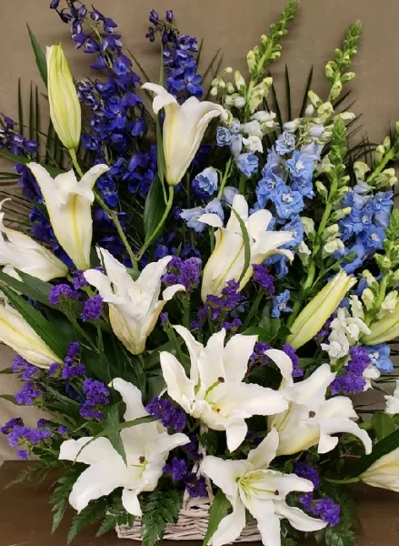 A close-up photograph of a large, intricate floral arrangement featuring white lilies, blue delphiniums, and greenery.