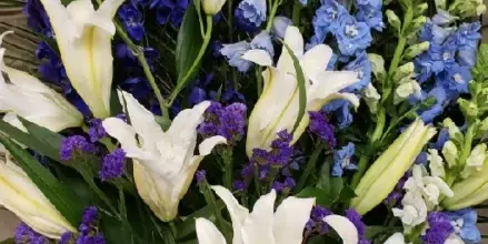 A close-up photograph of a large, intricate floral arrangement featuring white lilies, blue delphiniums, and greenery.