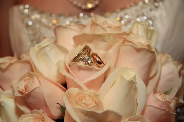 A close-up photograph of a wedding bouquet featuring cream-colored roses with a gold wedding ring and diamond engagement ring resting on the petals.