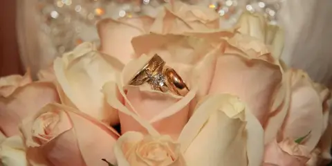 A close-up photograph of a wedding bouquet featuring cream-colored roses with a gold wedding ring and diamond engagement ring resting on the petals.