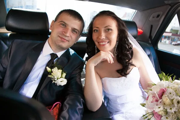 A smiling bride and groom sit together in the back seat of a black car service Newark. The groom wears a dark suit with a white boutonniere, while the bride holds a bouquet of flowers. Both look happy