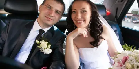 A smiling bride and groom sit together in the back seat of a black car service Newark. The groom wears a dark suit with a white boutonniere, while the bride holds a bouquet of flowers. Both look happy