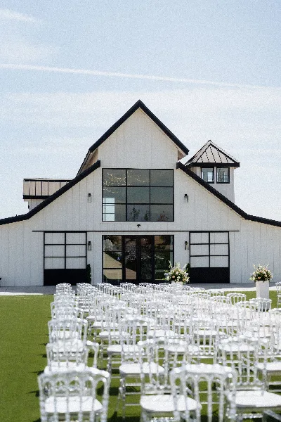 Exterior view of a white barn-style wedding venue with rows of white chairs set up on a green lawn for a ceremony.