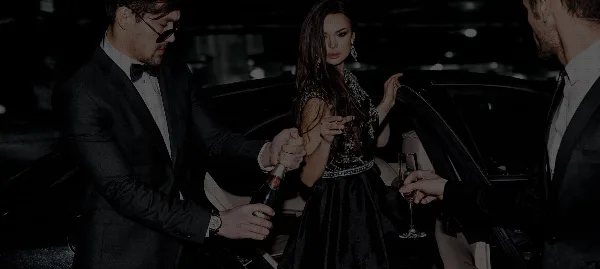 A professional chauffeur in a tuxedo assists a woman in an evening gown out of a luxury car.