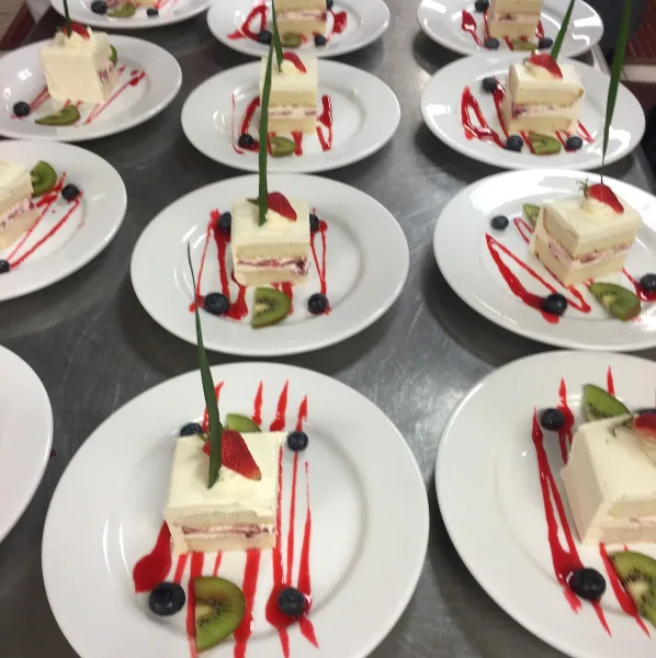 Multiple white plates featuring layered cake slices garnished with strawberries, kiwi, blueberries, and red syrup drizzle.