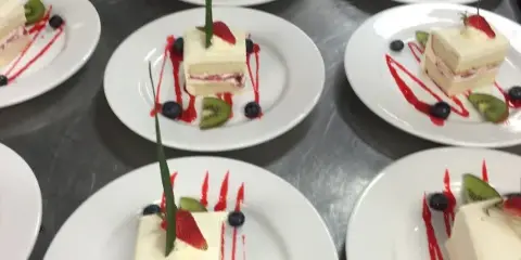 Multiple white plates featuring layered cake slices garnished with strawberries, kiwi, blueberries, and red syrup drizzle.