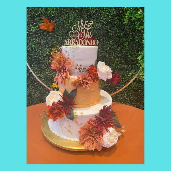 A three-tiered wedding cake with white and gold tiers, decorated with orange and red flowers and a Mr & Mrs topper.