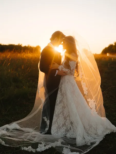 A bride and groom embracing in a field at sunset, with the bride's long lace veil flowing in the wind.