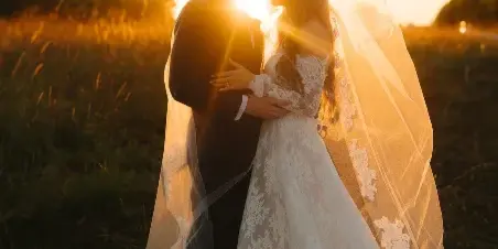 A bride and groom embracing in a field at sunset, with the bride's long lace veil flowing in the wind.