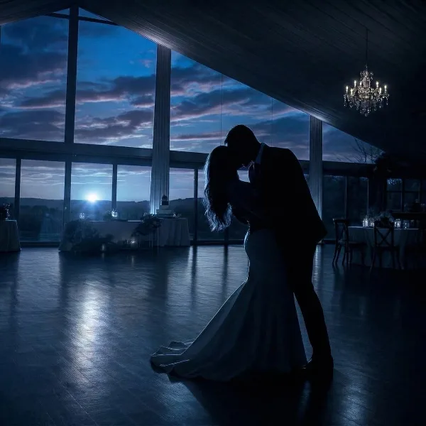 Silhouetted bride and groom dancing in a wedding reception hall with large windows and a chandelier at dusk.