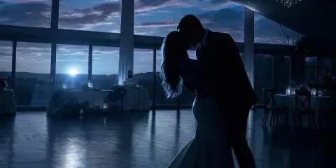 Silhouetted bride and groom dancing in a wedding reception hall with large windows and a chandelier at dusk.
