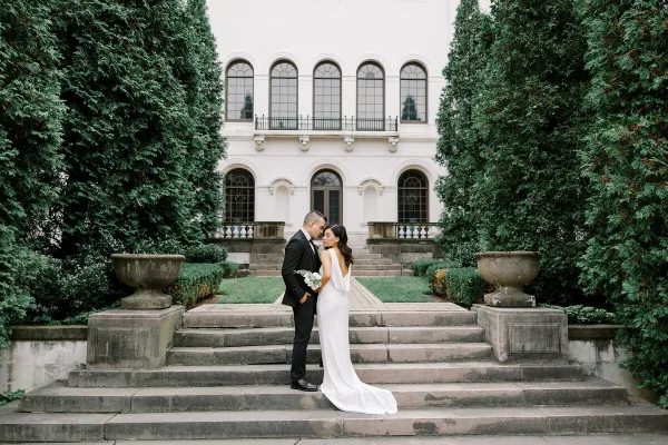 A bride and groom standing on stone steps in front of a historic white building with arched windows and manicured greenery.