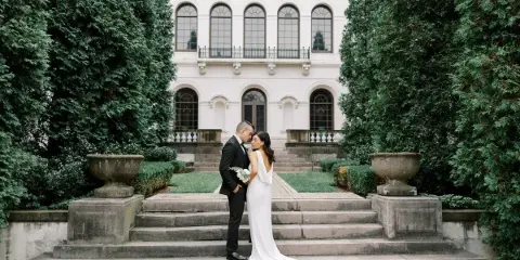 A bride and groom standing on stone steps in front of a historic white building with arched windows and manicured greenery.