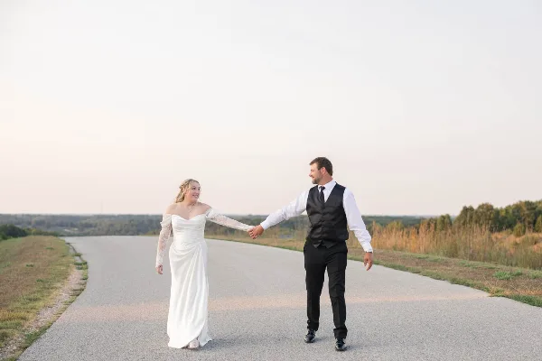 A bride and groom holding hands and walking down a paved road in a rural landscape.