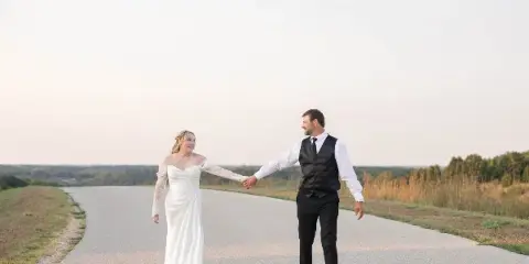 A bride and groom holding hands and walking down a paved road in a rural landscape.