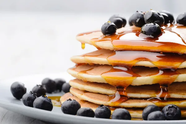 A tall stack of fluffy pancakes topped with fresh blueberries and drizzled with maple syrup on a white plate.