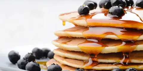 A tall stack of fluffy pancakes topped with fresh blueberries and drizzled with maple syrup on a white plate.