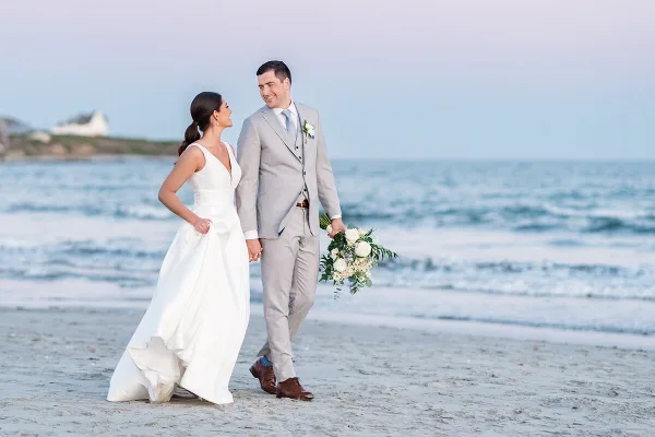 A bride and groom walking hand-in-hand on a sandy beach with the ocean in the background.
