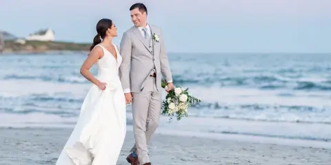 A bride and groom walking hand-in-hand on a sandy beach with the ocean in the background.