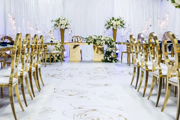 A wedding reception setup featuring gold chairs, white floral arrangements, and a custom aisle runner.