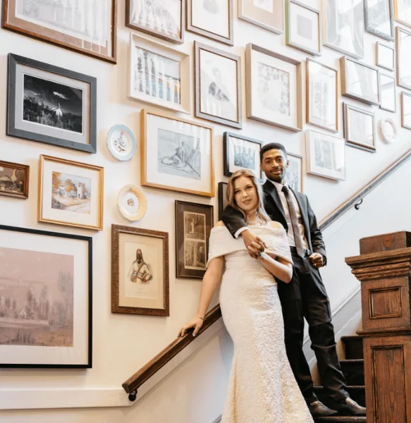 Bride and groom posing on a staircase in a venue interior with a wall of framed artwork.
