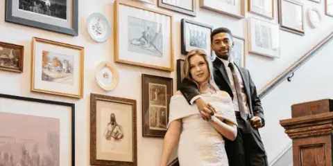 Bride and groom posing on a staircase in a venue interior with a wall of framed artwork.