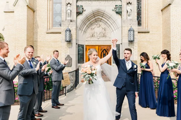 A bride and groom walk out of a church with their wedding party, raising their hands in celebration.