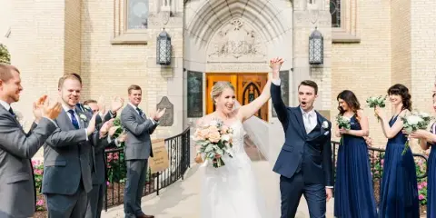 A bride and groom walk out of a church with their wedding party, raising their hands in celebration.