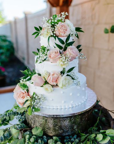 A three-tier white wedding cake decorated with pink and white roses and greenery, displayed on a silver stand.