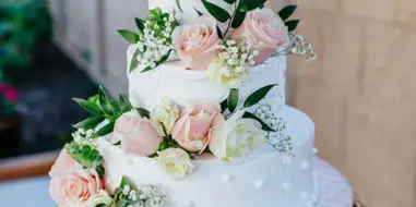 A three-tier white wedding cake decorated with pink and white roses and greenery, displayed on a silver stand.