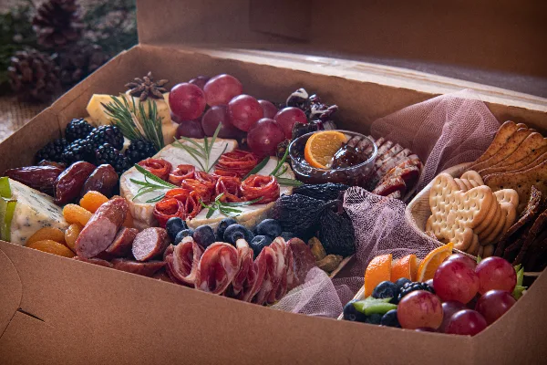A close-up of a curated grazing box containing assorted cheeses, rolled cured meats, grapes, blackberries, dried figs, and crackers.