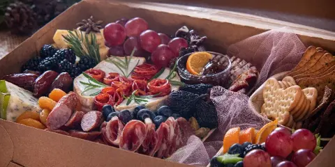 A close-up of a curated grazing box containing assorted cheeses, rolled cured meats, grapes, blackberries, dried figs, and crackers.