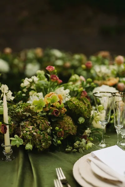 A close-up of a lush floral centerpiece featuring green hydrangeas, white and peach roses, and greenery, arranged on a table with a green tablecloth, candle, and glassware.