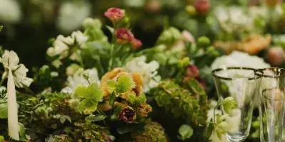 A close-up of a lush floral centerpiece featuring green hydrangeas, white and peach roses, and greenery, arranged on a table with a green tablecloth, candle, and glassware.