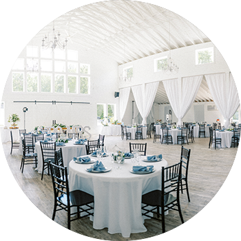 A spacious white wedding venue interior featuring round tables set with white linens, blue napkins, and floral centerpieces.