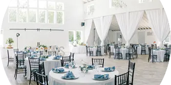A spacious white wedding venue interior featuring round tables set with white linens, blue napkins, and floral centerpieces.