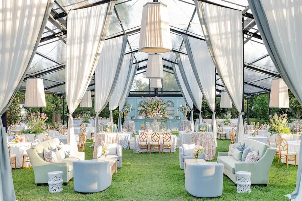 Luxury outdoor wedding reception inside a clear top tent with white drapery, floral centerpieces, and lounge seating.