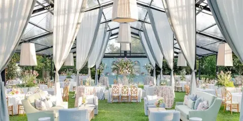Luxury outdoor wedding reception inside a clear top tent with white drapery, floral centerpieces, and lounge seating.
