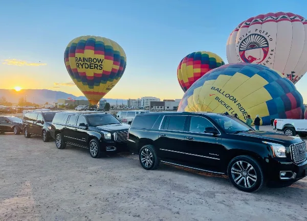 A row of black luxury SUVs parked on a dirt lot with hot air balloons in the background.