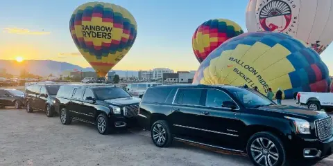 A row of black luxury SUVs parked on a dirt lot with hot air balloons in the background.