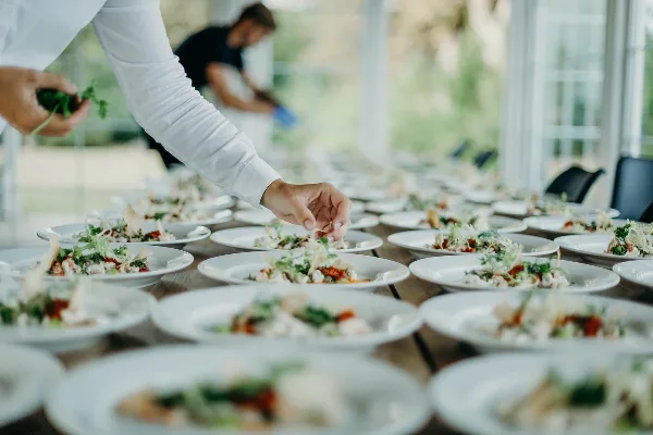A catering staff member in a white shirt plating food on a long table.