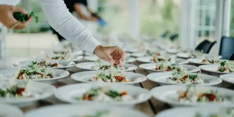 A catering staff member in a white shirt plating food on a long table.